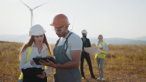 Equipo internacional de socios comerciales que se unen entre turbinas eólicas y examinan los planos. Grupo de profesionales que cooperan para producir energía limpia y respetuosa con el medio ambiente. Energía eléctrica ecológica. — Vídeo de stock