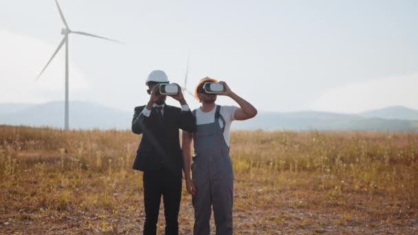 Afrykański amerykański biznesmen i indyjski inżynier noszący słuchawki VR podczas spotkania na farmie z turbinami wiatrowymi. Innowacyjne technologie i koncepcja alternatywnej energii. Dwóch partnerów korzystających z zestawu słuchawkowego VR — Wideo stockowe