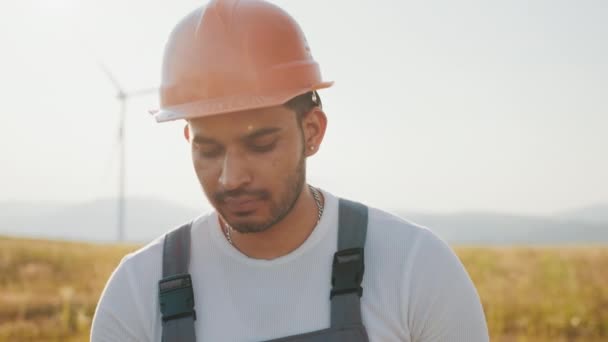 Técnico profesional en casco y overoles sonriendo oj cámara mientras está de pie en eco granja con aerogeneradores. Hombre indio usando tableta digital mientras controla el proceso de fabricación de energía limpia. — Vídeos de Stock