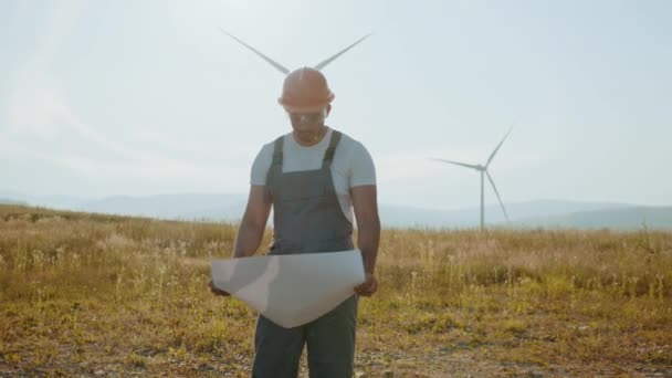 Indiase technicus in overalls, helm en bril controleren blauwdrukken van windmolens tijdens het staan op het platteland. Bevoegde industriearbeider die het proces van groene energieproductie controleert. Windmolen — Stockvideo