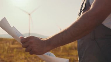 İş tulumlu, miğferli ve gözlüklü Hintli teknisyen kırsal bölgede dururken yel değirmenlerinin planlarını kontrol ediyor. Endüstriyel işçiler yeşil enerji üretim sürecini kontrol ediyorlar.