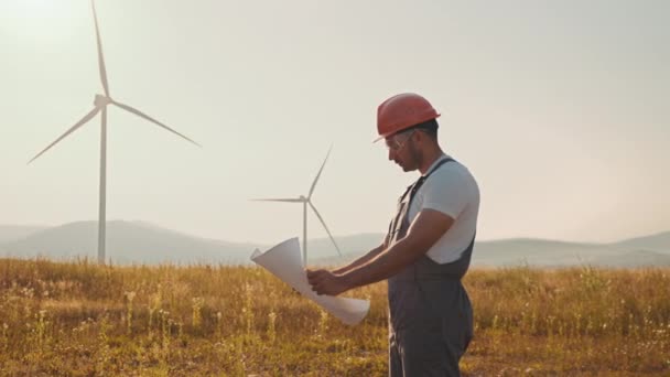 Indiase technicus in overalls, helm en bril controleren blauwdrukken van windmolens tijdens het staan op het platteland. Industriële werknemer controle proces van groene energieproductie. Eco-elektrisch vermogen. — Stockvideo