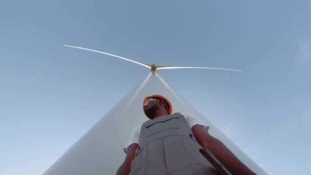 ヘルメット、眼鏡のアフリカ系アメリカ人技術者は、風力タービンでフィールドに立っている間、カメラを見て全体。安全性の電気と革新の概念。背景に風力タービン. — ストック動画