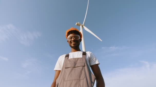 Técnico afro-americano em capacete, óculos e macacão olhando para a câmera enquanto estava em campo com turbinas eólicas. Conceito: energia renovável, tecnologia, eletricidade, serviço, verde, futuro. — Vídeo de Stock