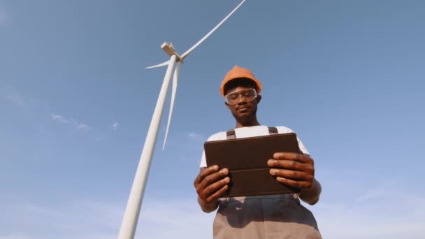 Kompetentny technik korzystający z tabletu cyfrowego podczas sterowania procesem pracy turbin wiatrowych na zewnątrz. Afrykański Amerykanin w szarym kombinezonie, pomarańczowym hełmie i okularach ochronnych. Turbiny wiatrowe — Wideo stockowe