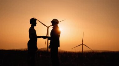 Siluetteki adam arka planda rüzgar türbinleri olan bir mühendisle tokalaşıyor. Çevre için temiz enerjinin iç açıcı görüntüsü. İki meslektaş başarılı işbirliği konusunda anlaşma yapıyor