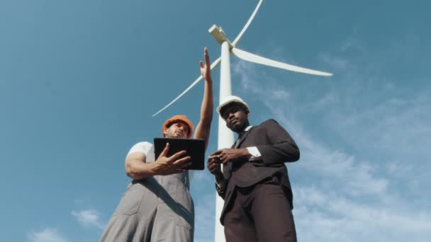 Multiraciala medarbetare undersöker vindkraftverk med hjälp av surfplatta. Medarbetare undersöker vindkraftverk med hjälp av moderna surfplattor utomhus. Två män i uniform och kostym talar om ren alternativ energi. — Stockvideo