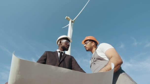 Inspector afroamericano con planos en las manos conversando con la concepción ingeniero indio entre granjas con turbinas eólicas. Energía limpia concepto de energía renovable, salvar el mundo. — Vídeos de Stock