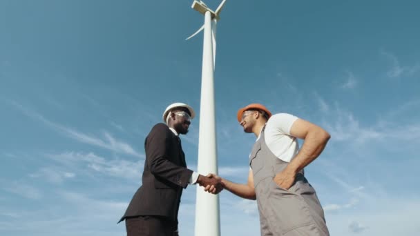 Африканський інспектор у білому шоломі та чорний костюм, тримаючи за руки інженера-індіана, стоячи біля величезного вітряка. Концепція: відновлювані джерела енергії, технології, електрика, послуги, зелений колір, майбутнє. — стокове відео