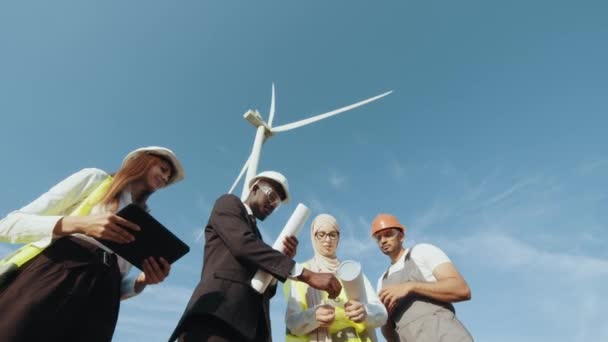 Cuatro socios multiétnicos en cascos de seguridad que estudian los planos de los aerogeneradores mientras están de pie juntos en el área rural. Trabajadores industriales competentes discutiendo proyectos comunes al aire libre. Energía limpia — Vídeo de stock