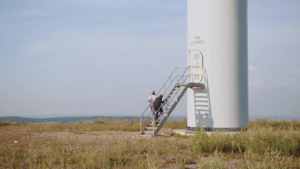 Africký americký inspektor v černém obleku a v bílé helmě mluví s indiánským technikem v uniformě při zkoumání větrné mlýnské farmy. Inženýr a podnikatel kráčející po poli s větrnými turbínami — Stock video