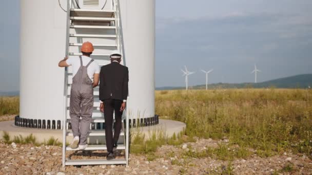 風力タービンとフィールドに一緒に歩いてインドのエンジニアとアフリカ系アメリカ人のビジネスマンのバックビュー。風車の仕事について技術者と話してた。二人の工業労働者 — ストック動画