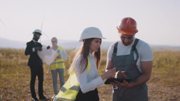 Tým multiraciálních průmyslových pracovníků v bezpečnostních helmách kontrolujících proces výroby zelené energie na farmě pomocí větrných turbín. Čtyři kolegové pomocí plánů a tabletu venku. Čistá energie — Stock video