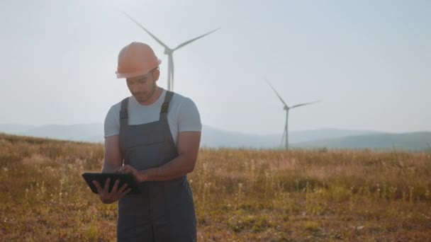 Portrét zaostřeného indiána stojícího na poli s větrnými turbínami a používajícího digitální tablet. Příslušný inženýr v oranžové helmě a šedé kombinéze. Muž v uniformě na statku s větrnými mlýny — Stock video