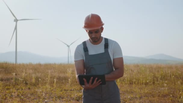 Porträtt av fokuserad indian man står på fältet med vindkraftverk och med hjälp av digitala tablett. Kompetent ingenjör med orange hjälm och grå overaller. Tekniker på lantgård med vindkraftverk. — Stockvideo