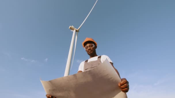 Afroamerikaner in Uniform, Helm und Brille arbeiten mit Blaupausen zwischen Windkraftanlagen. Professioneller Techniker überprüft und wartet die Produktion von grüner Energie. Hintergrund einer Windmühle — Stockvideo