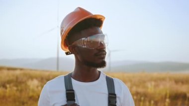 Yazın gün batımında elinde kocaman yel değirmenleriyle sahada dikilen Afrikalı Amerikalı adam portresi. Profesyonel teknisyen gri tulum, turuncu kask ve güvenlik gözlüğü takıyor. Alternatif enerjiyi kabul et