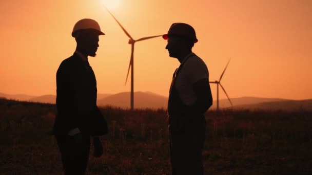 Silhouette eines afrikanisch-amerikanischen Ingenieurs und eines indischen Inspektors mit digitalem Tablet während eines Treffens im Freien und beim Händeschütteln. Zwei Partner stehen bei traumhaftem Sonnenuntergang auf einem Feld mit Windrädern. — Stockvideo