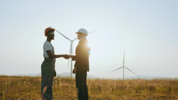 Dois colegas fizeram um acordo sobre uma cooperação bem sucedida. Conceito de pessoas, trabalho em equipe e energia ecológica. Técnico e inspetor afro-americano apertando as mãos na fazenda com turbinas eólicas. — Vídeo de Stock