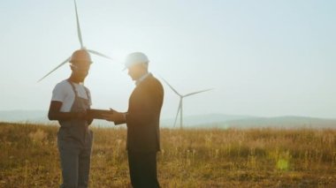 Modern tablet kullanarak rüzgar türbinlerini inceleyen iş arkadaşları. Mühendis ve müfettiş yel değirmeni çiftliğinde birlikte çalışıyorlar. Afro-Amerikan mühendis ve Hint müfettişi dışarıda dijital tablet kullanıyorlar.