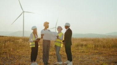 Uluslararası iş ortakları ekibi rüzgar türbinleri arasında bir arada duruyor ve planları inceliyor. Bir grup profesyonel, temiz çevre dostu enerji üretmek için işbirliği yapıyor. Rüzgar türbinleri