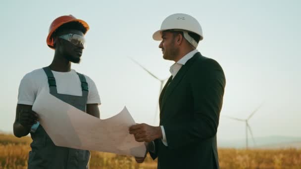 Primer plano dos técnicos profesionales en casco usando un gran plano. Ingenieros trabajando discutiendo en la estación ecológica. Central eólica. Planes de turbinas eólicas industriales. — Vídeo de stock