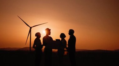 Silüet çoklu etnik mühendisler, teknisyenler ve güvenlik kasklı yöneticiler açık havada yeşil enerji hakkında toplantı yapıyorlar. Erkekler ve kadınlar yel değirmeni çiftliğinde çalışırken tablet ve çizim kullanıyorlar