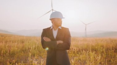 Siyah takım elbiseli Hintli işadamı, beyaz miğfer ve güvenlik gözlükleri. Rüzgâr türbinleriyle tarlada çapraz kollarla poz veriyorlar. Arka planda muhteşem bir yaz günbatımı. Yel değirmeni çiftliğinde poz veren Hintli adam