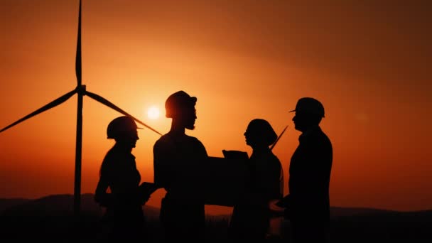 Men and women using tablet and drawing while working on windmill farm. Silhouette Multi ethnic group of engineers, technicians and managers in safety helmets having meeting outdoors about green energy — Stock Video