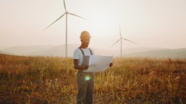 Elinde planları olan odaklanmış Afro-Amerikan bir adam rüzgar türbinleriyle çiftliğin arasında duruyor. Tulum, kask ve gözlük takan bir adam. Yeşil Enerji. Rüzgâr çiftliğinde çalışan üniformalı bir mühendis.