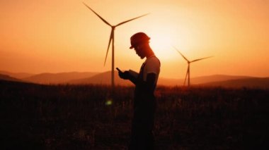 Siluet Afrikalı Amerikalı endüstriyel işçiler, rüzgar türbinleriyle çiftlikte dikilirken mobil konuşmalar yapıyorlar. Kasklı ve üniformalı bir mühendis eko istasyonunda çalışıyor. Rüzgar türbinleri olan bir adam.