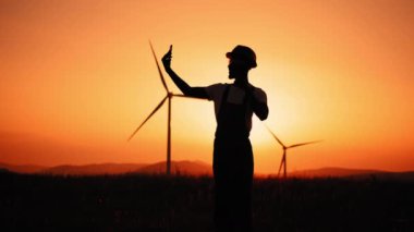 Siluet Pozitif Üniformalı ve miğferli mühendis modern akıllı telefonu yel değirmeni çiftliğinde görüntülü arama için kullanıyor. İnsanlar, teknoloji ve yeşil enerji kavramı. Eko istasyonunda çalışıyorum.