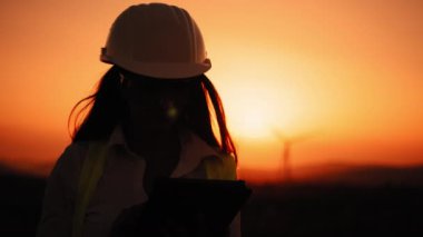 Siluet. Beyaz kasklı beyaz kadın yenilenebilir enerji çiftliğinde dijital tabletle çalışıyor. Dışarıdaki rüzgar türbinlerini kontrol eden kadın müfettiş. Yel değirmeni çiftliğinde çalış