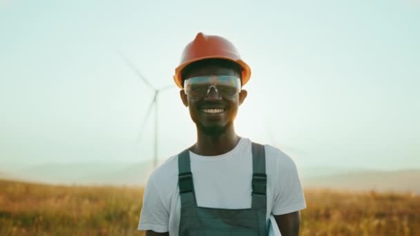 Afrikansk amerikan står på fältet med enorma väderkvarnar under sommarsolnedgången. Professionell tekniker i grå overaller, orange hjälm och skyddsglasögon. Porträtt av en ingenjör vindkraftverk — Stockvideo