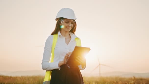 在可再生能源农场，戴着白色头盔的美丽的高加索女人与数字平板电脑一起工作。在室外控制风力涡轮机运行的女检查员。风车农场的工作 — 图库视频影像