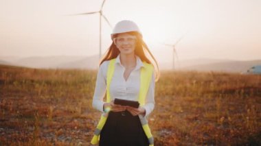 Yenilenebilir enerji çiftliğinde kasklı bir kadın tabletle çalışıyor. Yel değirmeni çiftliğinde resmi kıyafetler, miğfer ve gözlük takan kadın. İnsan kavramı, teknoloji ve yeşil enerji.