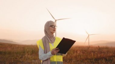 Türbanlı ve gözlüklü çekici Müslüman kadın yel değirmenleriyle çiftlikte dikilirken panodaki bazı belgelere bakıyor. Yaz batımı. İnsan kavramı, teknoloji ve yeşil enerji