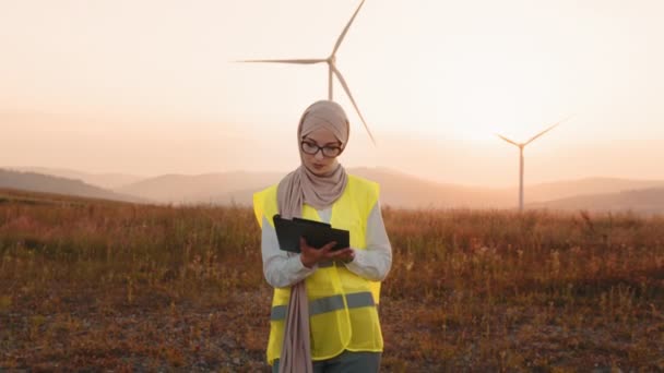Muslimka drží podložku, zatímco pózuje na větrném mlýně. Portrét inspektorky na ekologické farmě během letního západu slunce. Žena v hidžábu stojící na větrné farmě se schránkou — Stock video