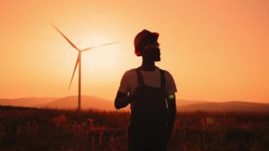 Siluet Afrikalı adam yaz günbatımında büyük yel değirmenleriyle sahada duruyor. Profesyonel teknisyen tulum, turuncu kask ve güvenlik gözlüğü takıyor. Bir mühendis rüzgar türbininin portresi