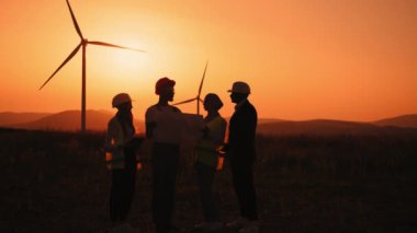 Silüet çoklu etnik mühendisler, teknisyenler ve güvenlik kasklı yöneticiler açık havada yeşil enerji hakkında toplantı yapıyorlar. Erkekler ve kadınlar yel değirmeni çiftliğinde çalışırken tablet ve çizim kullanıyorlar