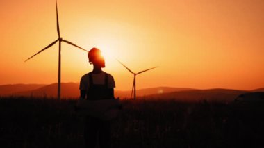 Siluet mühendisi sabit şapka standında kameraya bakıyor. Akşamları ekolojik istasyonun büyük bir planını tutuyor. Rüzgar türbinleri. Elinde planlar olan Afrikalı adam rüzgar türbinleri olan çiftlikler arasında