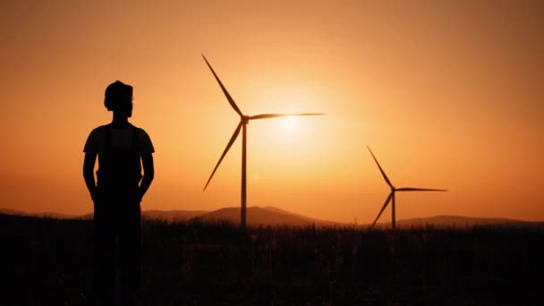 Силуэт человека, стоящего на поле с огромными ветряными мельницами во время летнего заката. Концепция загрязнения окружающей среды, новые технологии альтернативной энергетики. Фон мельницы. — стоковое видео