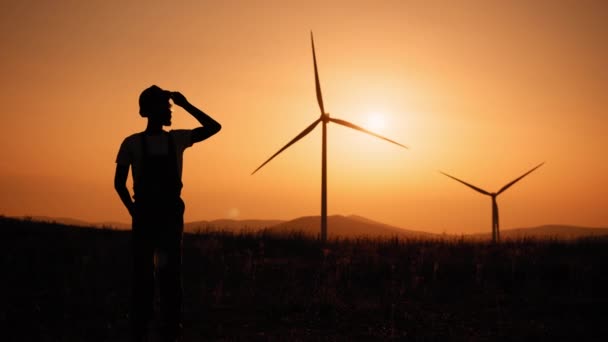 Silhuett Cinematic skott av ung ingenjör sätta säkerhetshjälm på nöjd med sitt jobb på bakgrund av vindkraftverk med vid solnedgången.Koncept: förnybar energi, teknik, el, service — Stockvideo