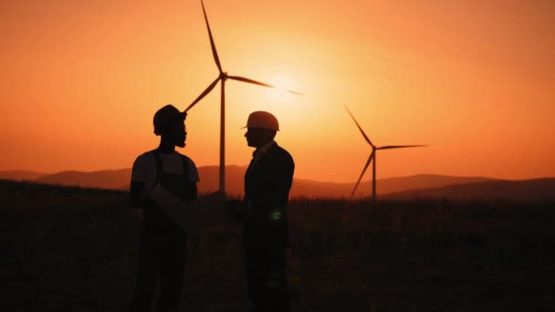 Silhouette Deux ouvriers industriels multiraciaux examinant les plans des éoliennes avec incroyable. Zone rurale avec éco-ferme de production propre énergie verte. Concept travail d'équipe et énergie respectueuse de l'environnement. — Video