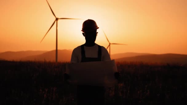 Silhouette uomo afroamericano con cianografie in mano in piedi tra fattoria con turbine eoliche. Ingegnere competente con tuta, casco arancione e occhiali di sicurezza. Ingegnere tra i mulini a vento — Video Stock