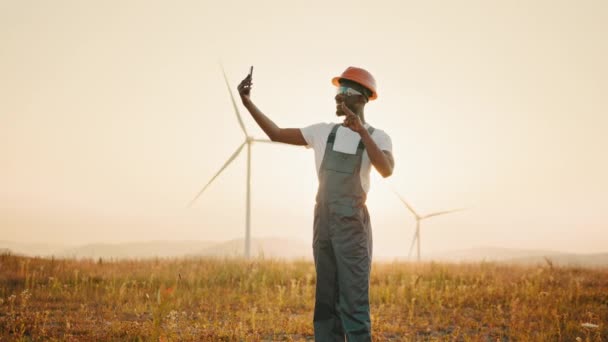 Ingénieur positif en uniforme et casque à l'aide d'un smartphone moderne pour l'appel vidéo parmi le parc éolien. Concept de personnes, de technologie et d'énergie verte. Travailler sur une éco station — Video