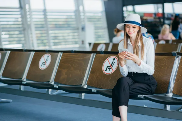 En ung kvinne med ansiktsmaske på flyplassen bruker smarttelefonen sin. Sender tekstmeldinger, ser på kamera, sjekker e-poster på internett, en vellykket kvinnelig leder på jobben – stockfoto