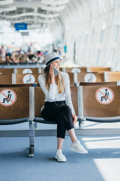 Ung kvinne på flyplassen med vogn bag, snakker i telefonen, ser på kamera og smiler . – stockfoto