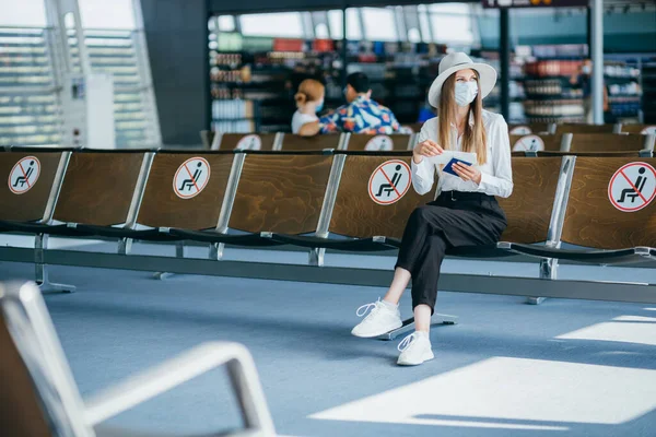 Maskierter Tourist sitzt in der Lounge des Flughafenterminals auf leeren, markierten Stühlen unter neuen, normalen und lebendigen sozialen Abstandsregelungen. Frau sitzt in Flughafen-Lounge und schaut auf ihren Reisepass — Stockfoto