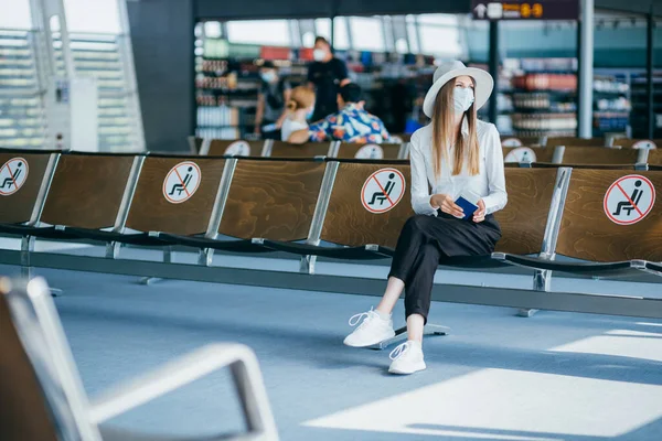 Turista mujer con máscara con billetes de avión y sentada en una silla socialmente distanciada en el aeropuerto durante la epidemia de coronavirus. Concepto de nueva normalidad del transporte aéreo debido a la pandemia —  Fotos de Stock